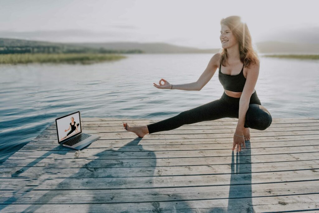 Frau macht Online Yoga und sitzt mit Laptop an einem See